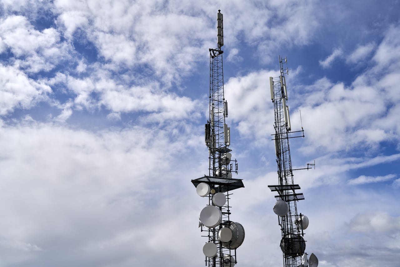Telecommunication towers with antennas and dishes under a bright cloudy sky.